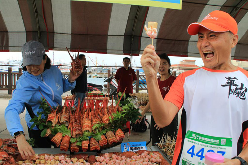 2025 菊島澎湖跨海馬拉松特色「痛風補給站」,桌上擺滿整隻豪華龍蝦,跑者興奮地拿著龍蝦串大快朵頤,表情驚喜。