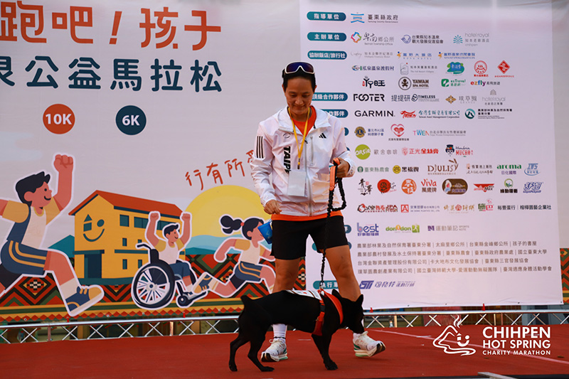 三鐵一姐李筱瑜帶著愛犬『金教練』,溫馨出席 2025 跑吧!孩子知本溫泉公益馬拉松,背景可見活動主視覺與品牌合作夥伴 正光製藥 Logo。