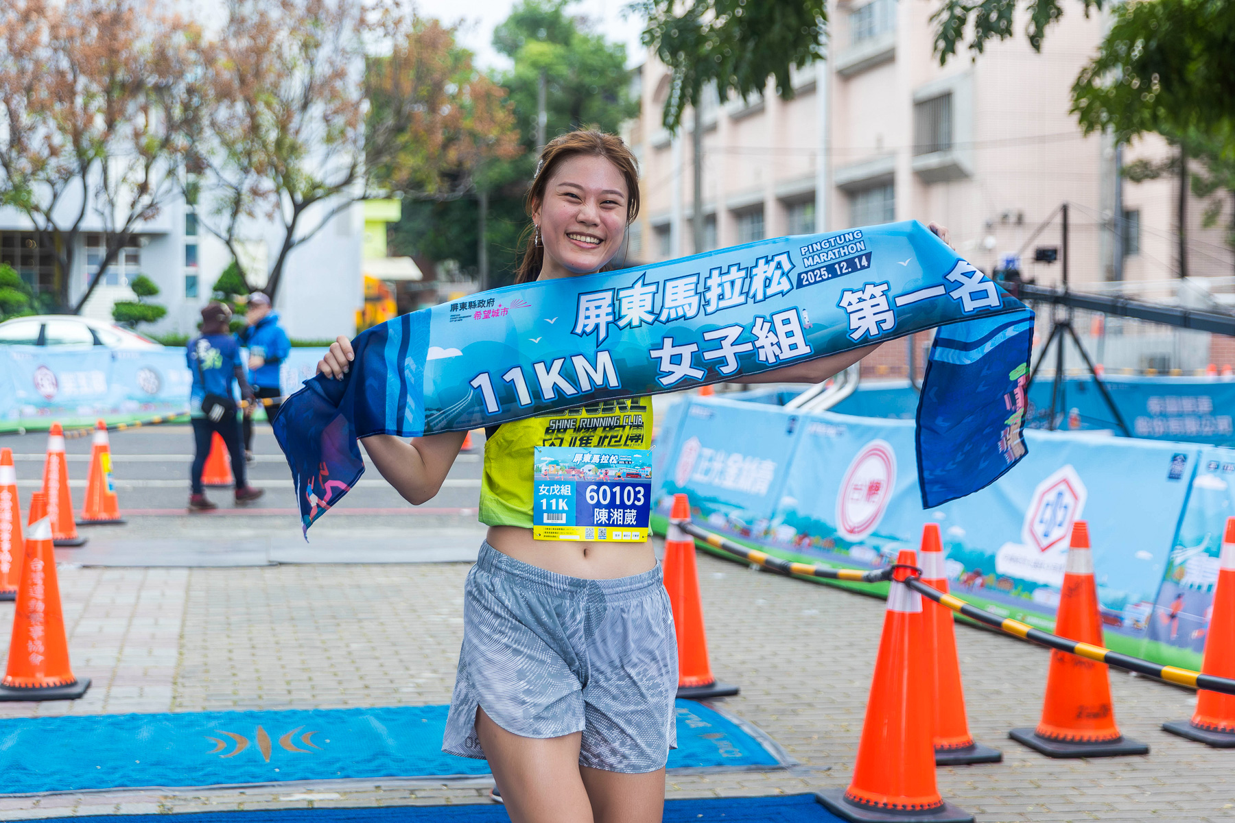 屏東馬拉松 11 公里女子組完賽畫面，跑者於終點開心舉起第一名紀念布條，展現完成比賽後的喜悅與賽事榮耀，現場動線與賽事佈置清楚。
