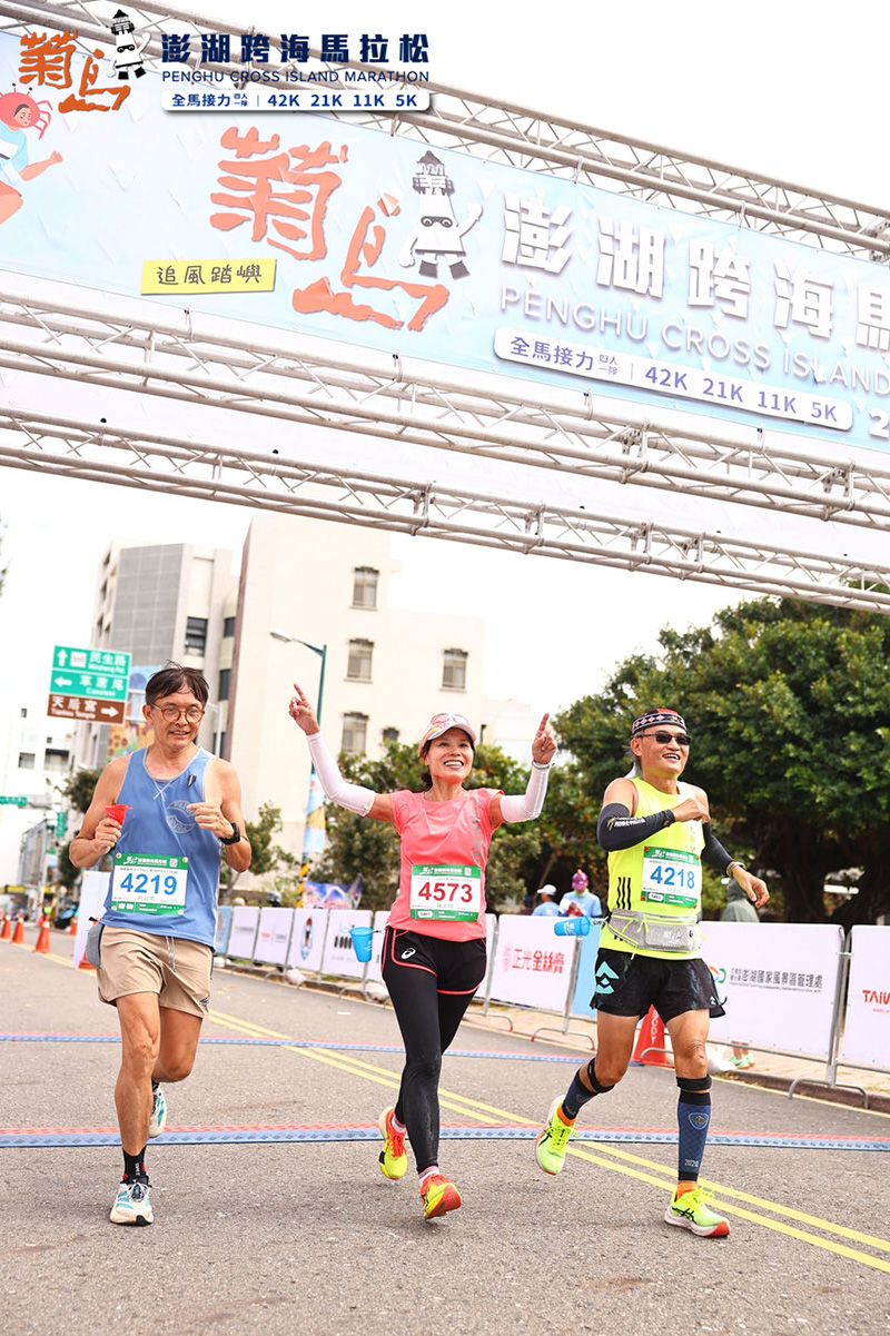 2025 菊島澎湖跨海馬拉松跑者抵達終點前的精彩瞬間，三位參賽者臉上掛著燦爛笑容，中間的女跑者高舉雙手慶祝，展現享受比賽與挑戰成功的快樂氛圍。