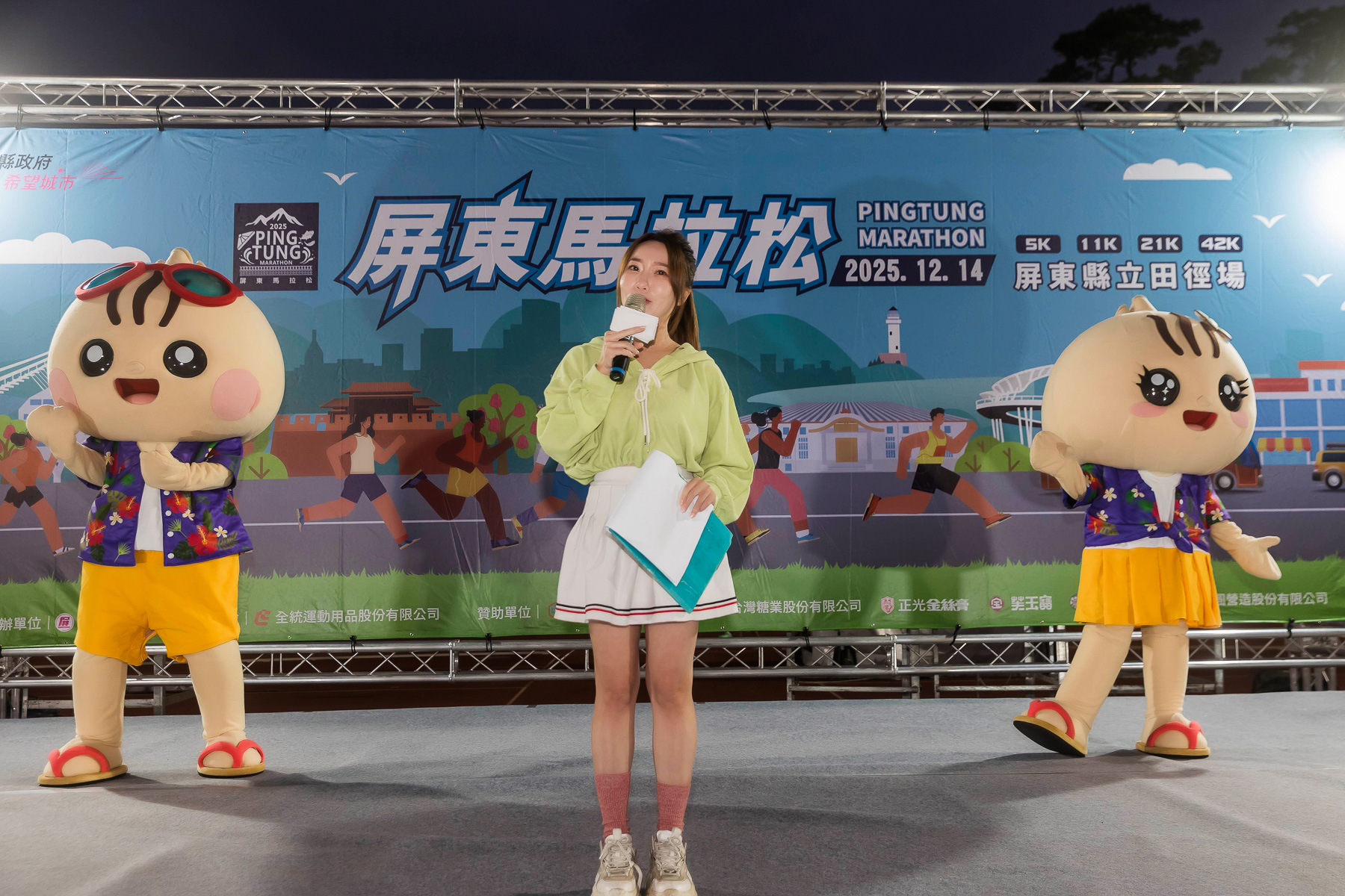 屏東馬拉松活動舞台畫面，主持人於舞台中央介紹賽事，兩側有吉祥物互動演出，背景為 2025 屏東馬拉松主視覺與賽事資訊，現場氣氛熱鬧歡樂。