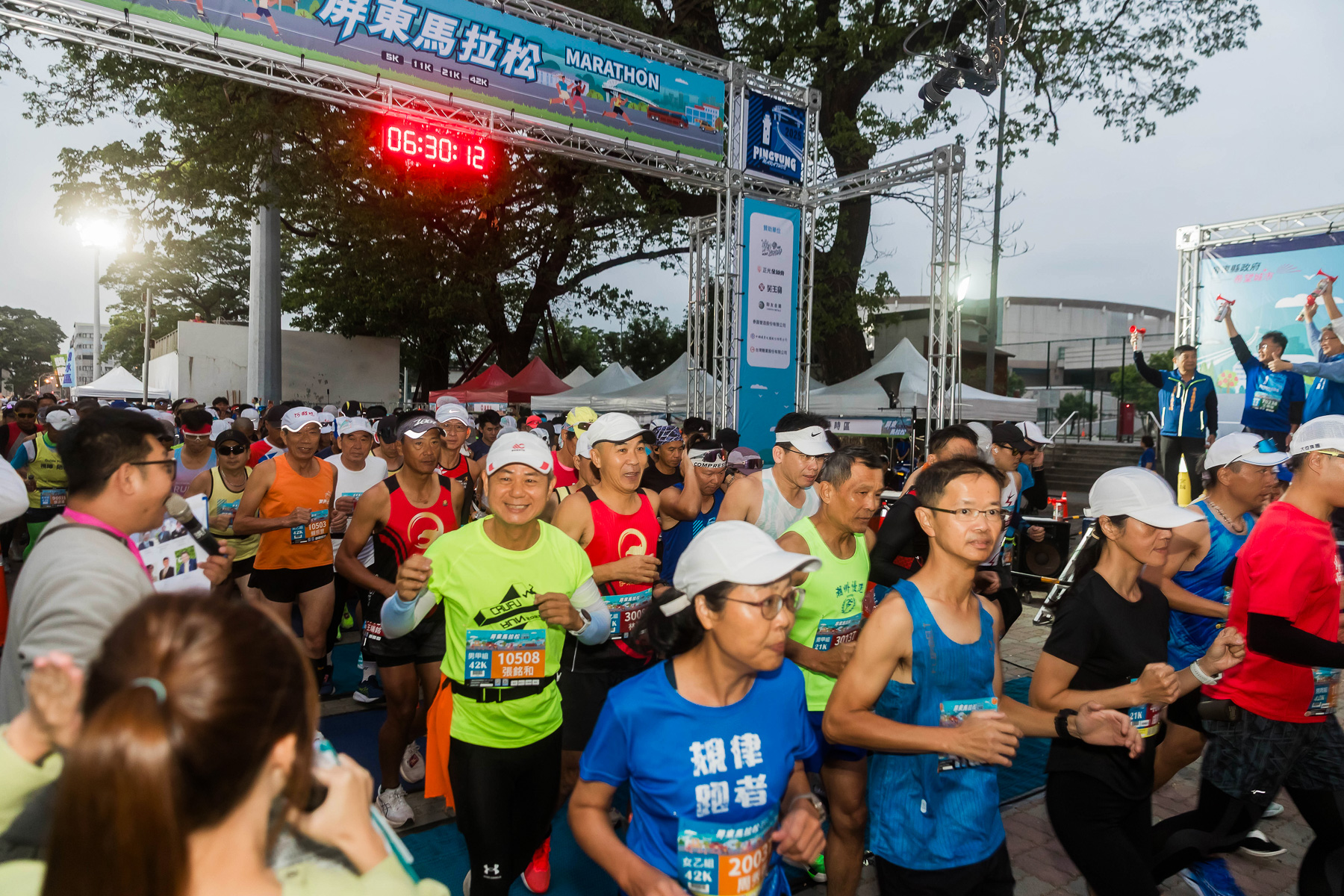 屏東馬拉松清晨起跑畫面，跑者從起跑拱門通過，穿著各色賽衣奔跑於賽道上，展現南台灣路跑賽事熱鬧且充滿活力的氛圍。