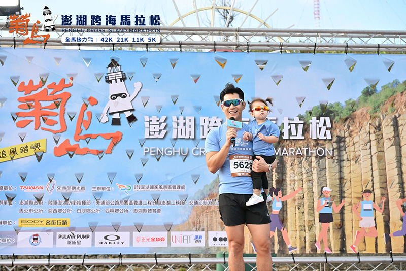 2025 菊島澎湖跨海馬拉松代言人健身男神祖雄(Hero Tai)抱著戴墨鏡的女兒開心出席活動,背景為活動主舞台與贊助商背板。